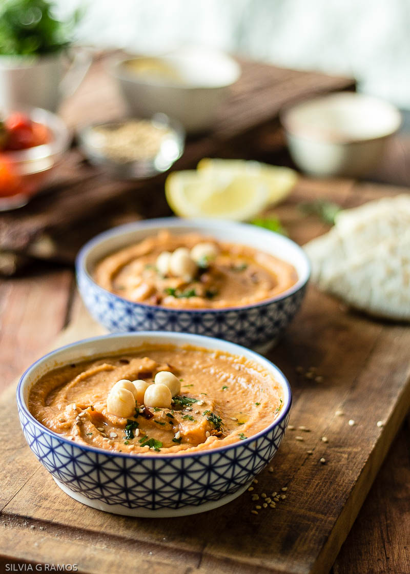 Ingredientes para hummus de pimiento rojo vegano: garbanzos, tahini, pimiento asado y especias frescas en tabla de madera rústica