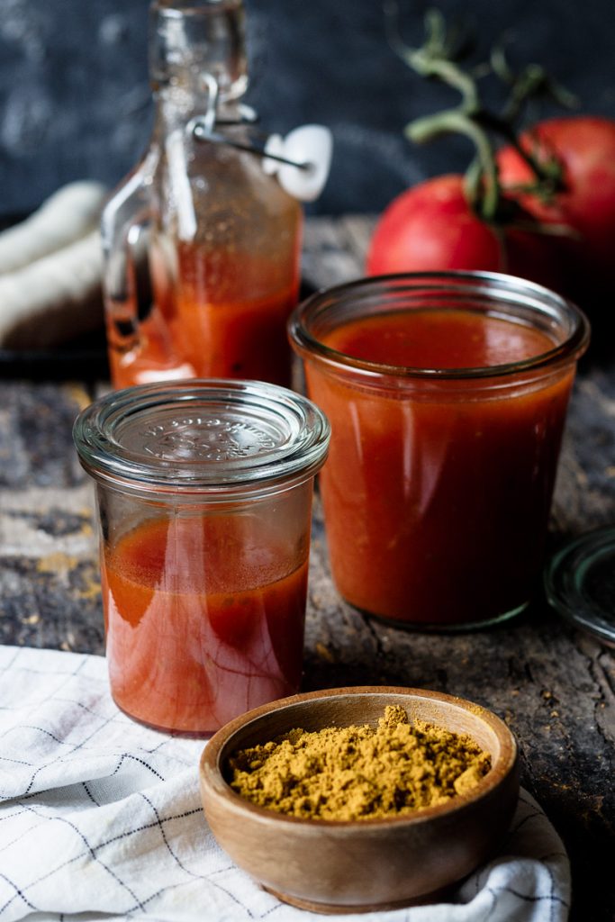 Tarros de ketchup de curry casero con textura suave y color rojizo, acompañados de curry en polvo y tomates frescos sobre una mesa rústica.